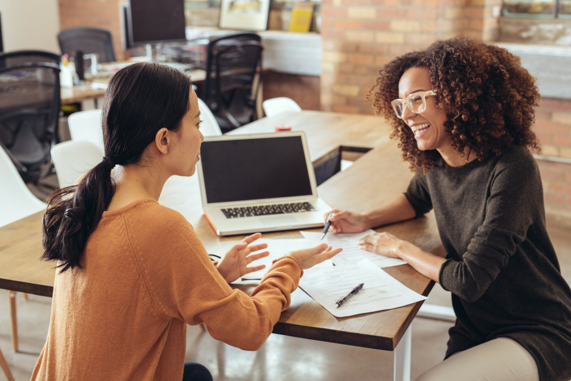 Two women smile and laugh while they work at a table with a laptop.